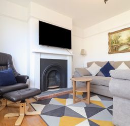 A living room with a beige sofa with cushions a black chair with footrest a fireplace with a TV mounted above a small wooden table and a geometric patterned rug at The Heights in Truro
