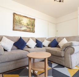 A living room with a beige sectional sofa with white and blue cushions a round wooden coffee table a geometric patterned rug and a framed landscape painting on the wall at The Heights in Truro