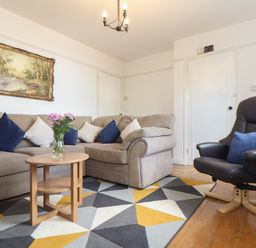 A living room with a beige sectional sofa with cushions, a wooden coffee table with flowers, a black armchair with a cushion, and a geometric patterned rug at The Heights in Truro