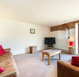 A living room with two brown sofas with red cushions a wooden coffee table a TV on a stand and a side table with a red lamp at Widgeon in Wheatacre