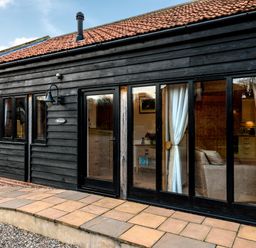 A black wooden exterior wall with windows and a glass door showing a living area at Pheasant in Wheatacre