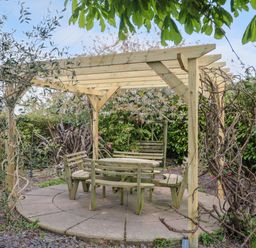 A wooden pergola over a circular paved area with wooden benches and a table in a garden at Te Papa in North Petherton