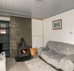 A living room with a sofa covered by a grey cover a wood stove with fire a tv on a white cabinet and a bookshelf at Curragh Cottage in Rathimney near New Ross County Wexford