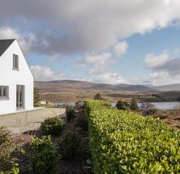 A white house with a dark roof beside a gravel path and green hedge with hills and a lake in the background at Moorhen House in Loughanure near Annagry County Donegal