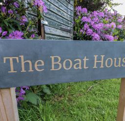 A wooden sign with the text The Boat House on a lawn with purple flowers and a green shed in the background at The Boathouse in Belford