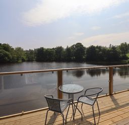 A wooden deck with a glass round table and two chairs overlooking a lake with trees at Otters Cove in Belford