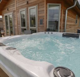 An outdoor hot tub with bubbling water next to a wooden house with windows and outdoor furniture at 4 Meadow Retreat in Dobwalls