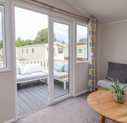 A living room with a sofa a wooden table and a plant next to glass doors leading to an outdoor seating area at Windermere Lodge in Morecambe