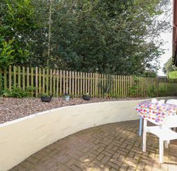 An outdoor patio area with a small table covered by a checkered tablecloth and two white chairs next to a house with a wooden fence and plants in the background at Paws-A-While in Hoveton & Wroxham
