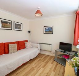 A living room with a red sofa, a small TV on a stand, framed pictures on the walls, and flowers on a table at Paws-A-While in Hoveton & Wroxham