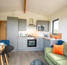 A kitchen with grey cabinets and appliances next to a green sofa with an orange pillow at Cae Pren in New Mills near Llanfair Caereinion