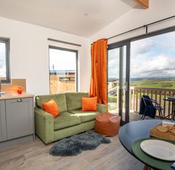 A living room with a green sofa and orange cushions next to a kitchen sink and large glass doors opening to a balcony with countryside view at Cae Pren in New Mills near Llanfair Caereinion