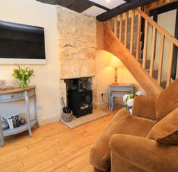 A living room with a brown sofa a stone fireplace a wood burning stove a wall mounted television a wooden staircase a console table with flowers and books at Broadway Cottage in Broadway