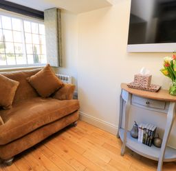 A brown couch with two cushions next to a window and a small blue table with books flowers and tissue box in a room at Broadway Cottage in Broadway