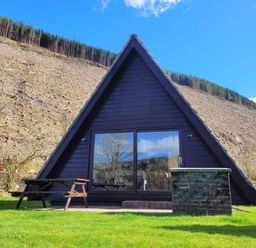 An a frame cabin with glass doors next to a wooden picnic table on grass with a cleared hillside in the background at Lodge 2 - Inglewood Lodge in Spean Bridge