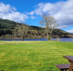 A grassy area with trees near a lake and a wooden picnic table at Lodge 2 - Inglewood Lodge in Spean Bridge
