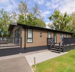 A single story wooden cabin with a deck and stairs surrounded by grass and trees at 18 Manleigh Park in Combe Martin