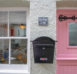 A window and a pink door with a black mailbox and a sign saying Sea Pinks c 1780 on a white textured wall at Sea Pinks in Looe