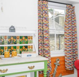 A room corner with a green and white cabinet with yellow kettle and cups next to a window with fish patterned curtains at Sea Pinks in Looe