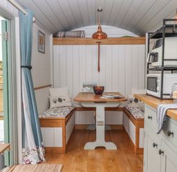 A dining area with a wooden table and bench seats with cushions inside a hut with a kitchen counter and appliances on the right at Burr 2 Shepherds Hut Swimbridge near Barnstaple