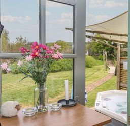 A table with flowers, a candle, and a sheep ornament by a window overlooking a garden and a hot tub at Burr 2 Shepherds Hut Swimbridge near Barnstaple