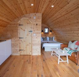 An interior wooden pod with a bed, a brown leather sofa, a coffee table, and a kitchen area with cabinets at Glamping Pod 1 Comfort in Lebberston