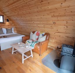 A wooden room with a bed a leather sofa a small wooden table and a wood-burning stove at Glamping Pod 1 Comfort in Lebberston