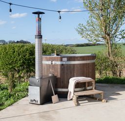 An outdoor hot tub with a wood stove heater on a concrete patio with a wooden step stool and string lights near trees and fields at Glamping Pod 1 Comfort in Lebberston