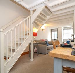 A living room with a staircase, gray sofa, side table with lamp, dining table set with plates and glasses at Cobblestone Cottage in Bude