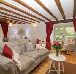 A living room with a grey sofa, red curtains, a rocking chair, and a small white table with flowers at 4 Barton Cottages Down Thomas near Plymouth