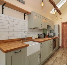 A kitchen with wooden countertops, green cabinets, a white farmhouse sink, toaster, microwave, and wooden door at The Cart Shed in Barwick near Yeovil Somerset