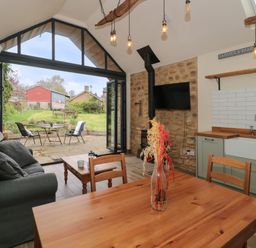 An open plan kitchen and living area with wooden dining table and chairs a gray sofa wood burning stove and glass doors opening to a patio at The Cart Shed in Barwick near Yeovil Somerset