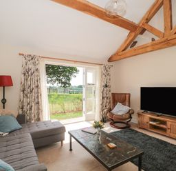 A living room with a gray sectional sofa a brown leather armchair a flat screen tv on a wooden stand a table with flowers and open doors showing a garden at Orchard View in Redmary D Abitot near Hartpury