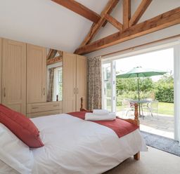A bedroom with a bed white bedding and red pillows wooden wardrobes exposed wooden beams and a patio door leading to a garden with a table chairs and a green umbrella at Orchard View Redmary D Abitot near Hartpury