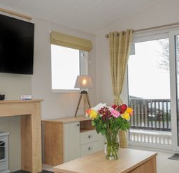 A living room with a television fireplace lamp flowers on a table and a sliding glass door at Lodge 74 at Riviera Bay Coastal Retreat in Brixham