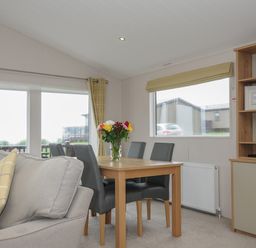 A dining area with a wooden table and four chairs next to a gray sofa with yellow pillows and a cabinet with shelves at Lodge 74 at Riviera Bay Coastal Retreat in Brixham