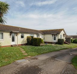 A row of single-story houses with tiled roofs and green lawns along a paved pathway at Chalet 7 at Landscove Holiday Park in Brixham