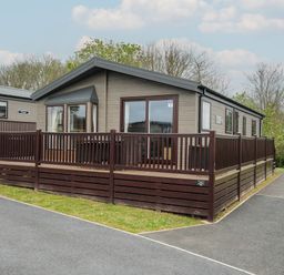 A single story cabin with a wooden deck and railing surrounded by pavement and grass at Lodge 72 at Riviera Bay Coastal Retreat in Brixham