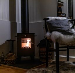A small wood-burning stove with fire inside and a chair with a fur throw in a corner of a room at La'al Ling in Penrith