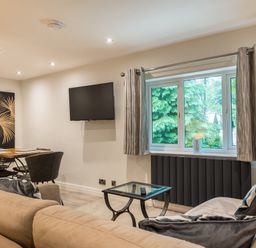 A living room with a beige sofa a glass coffee table a wall mounted TV a dining table with chairs and a window with curtains at Ullswater View Apartment in Ullswater