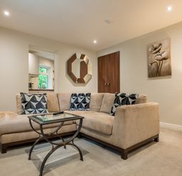 A living room with a beige sectional sofa black and white patterned cushions a glass coffee table a wall mirror a painting and a lamp at Ullswater View Apartment in Ullswater