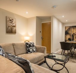 A living room with beige sofas and patterned cushions a lamp a glass coffee table and a dining table with chairs and artwork on the walls at Ullswater View Apartment Ullswater