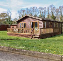 A wooden cabin with a railing deck and stairs surrounded by grass and trees at 3 The Glade in St Minver Rock