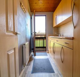 A narrow kitchen with wooden cabinets a sink two grey mats a white door and an open exterior door at 3 The Glade in St Minver Rock