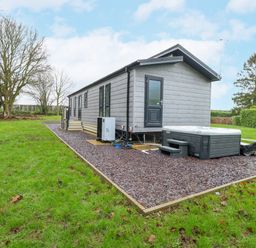 A small gray house with multiple windows and a door next to a hot tub on gravel surrounded by grass and trees at Wurzburc in Swaffham