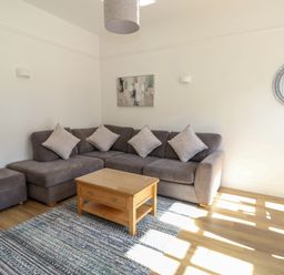 A living room with a grey sectional sofa and cushions a wooden coffee table a patterned rug a round mirror and wall art at Splash in Weymouth