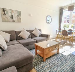 A living room with a grey sofa, a wooden coffee table, a glass dining table with chairs, and large windows at Splash in Weymouth