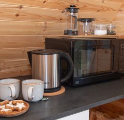 A kitchen counter with two mugs of tea a plate of star shaped cookies an electric kettle a microwave and glass containers on a wooden wall at Glamping Pod 3 Harmony Lebberston