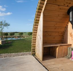 A wooden shelter with boots and a black stove pipe next to a grassy area with a pond at Glamping Pod 5 Shelter in Lebberston