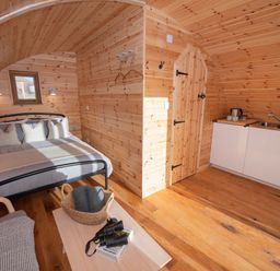 A wooden room with a bed pillows bedside lamps a small kitchen area a wooden table with a basket binoculars and books and a door at Glamping Pod 5 Shelter in Lebberston